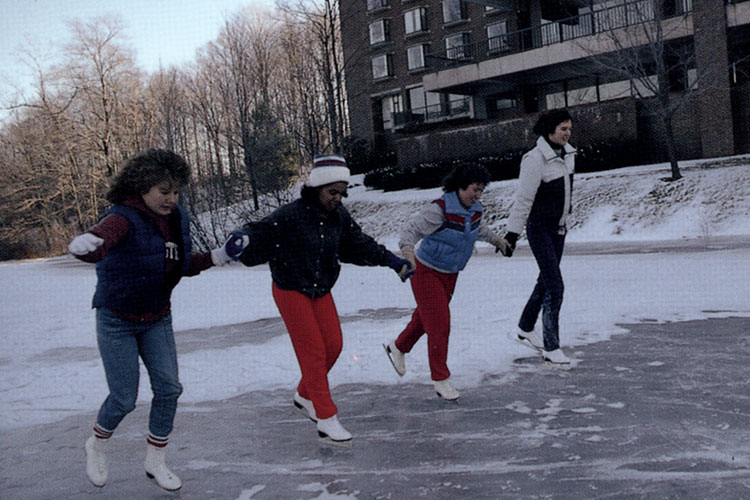 Four students ice skating on Upper Lake, with MacGregor Hall dormitory in the background