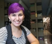 Yaine Neyhard sits in the library, wearing a striped shirt, with short, purple hair.