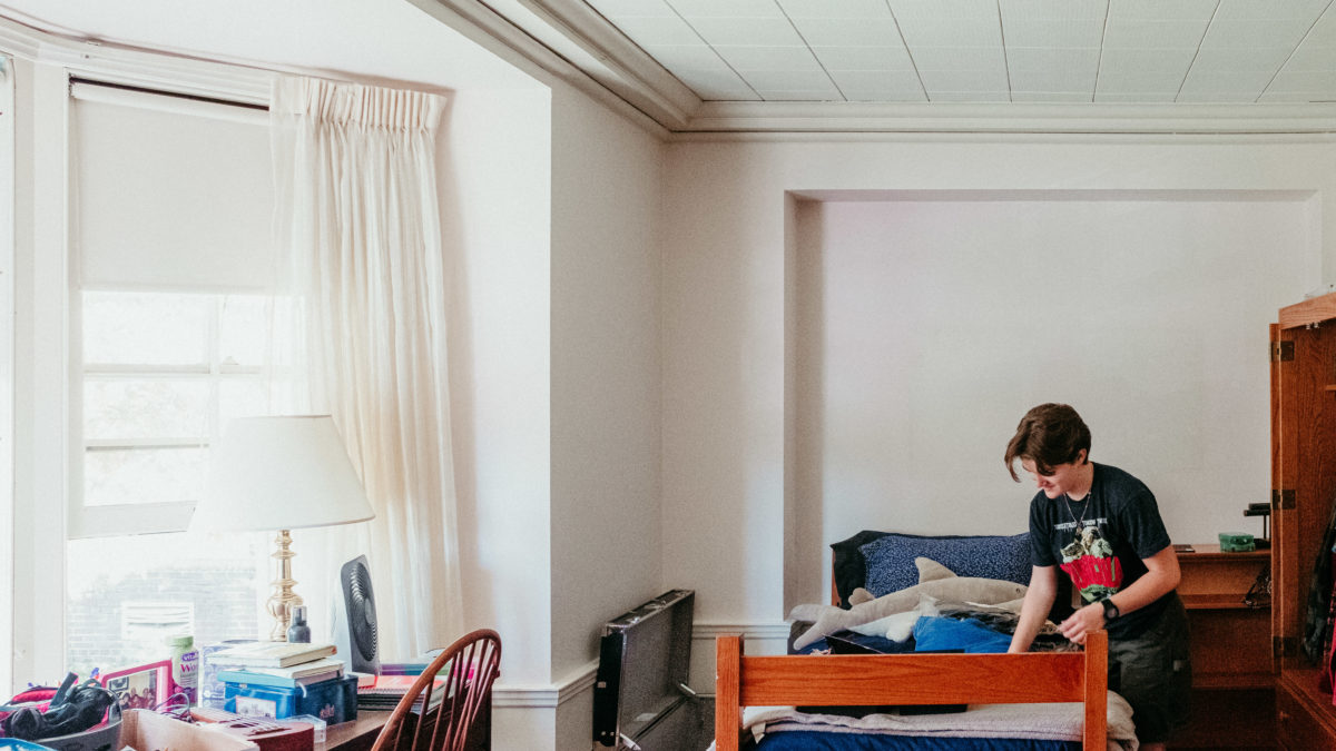 A student leans over their bed as they set up their room in Abbey Hall. Sunlight streams in from the large bay window to the left.