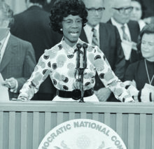 A black and white photo of Shirley Chisholm standing in front of a podium that reads Democratic National Convention. She is in the middle of speaking into the microphones, with her hands on the podium and leaning towards the audience.