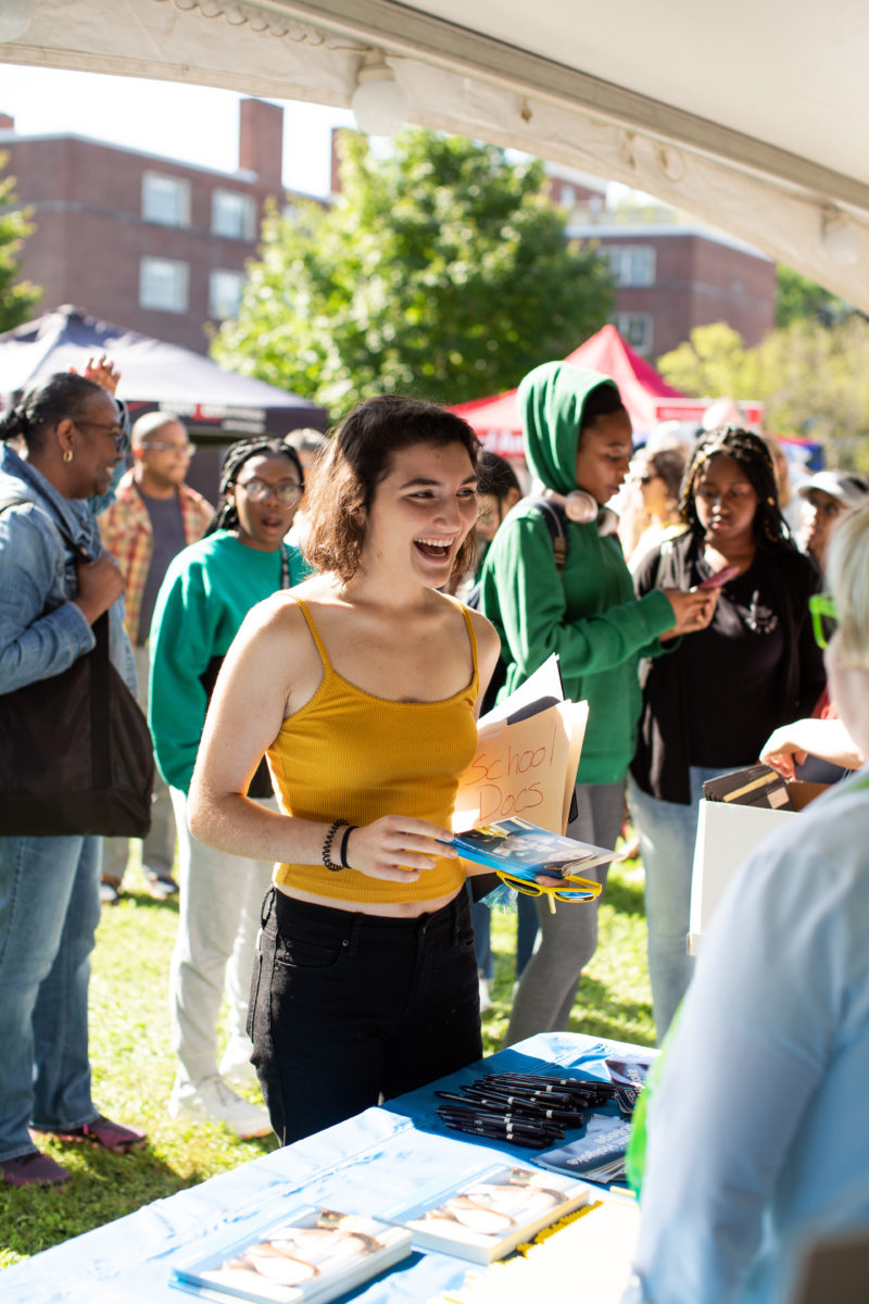 A student in a yellow tank top and black jeans smiles while speaking to people tabling at the Orientation welcome tent. There are people behind them walking to other tents and tables.