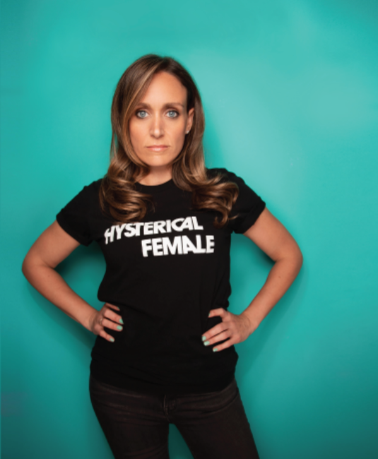 Jess Inserra '98 posing in front of a teal background. She stares straight into the camera with her hands on her hips and her shirt reads "hysterical female."